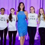 Maria stands on stage in blue dress with teens wearing girl power t-shirts