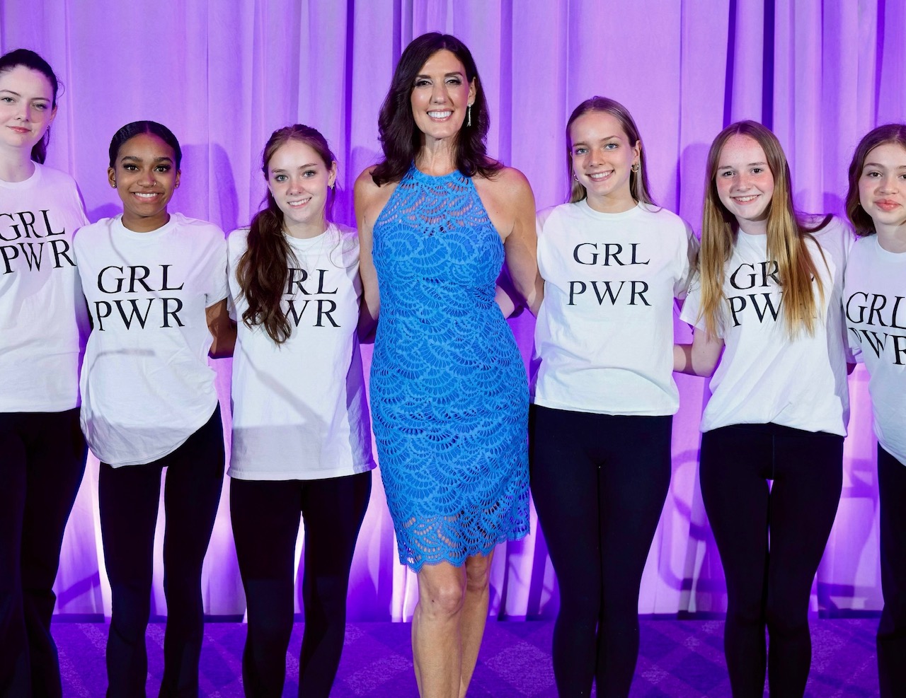 Maria stands on stage in blue dress with teens wearing girl power t-shirts